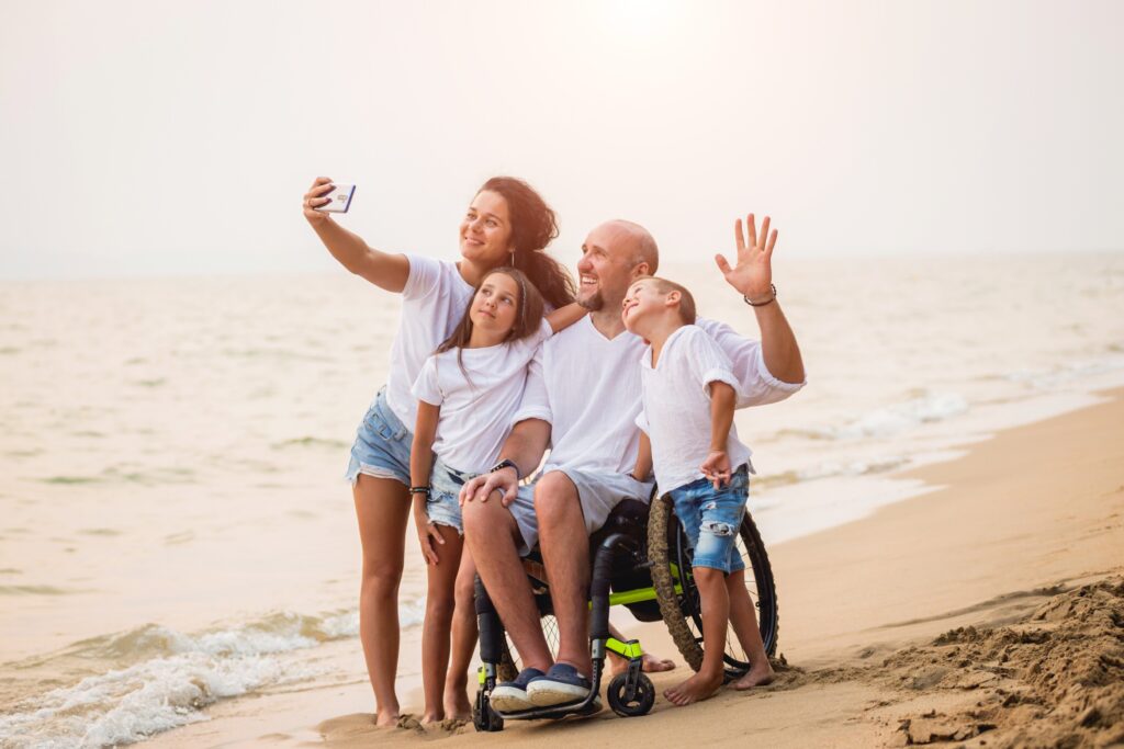 A family formed by two adults and two children are taking a selfie at the beach. The father is a wheelchair user. They're all smiling at the camera.