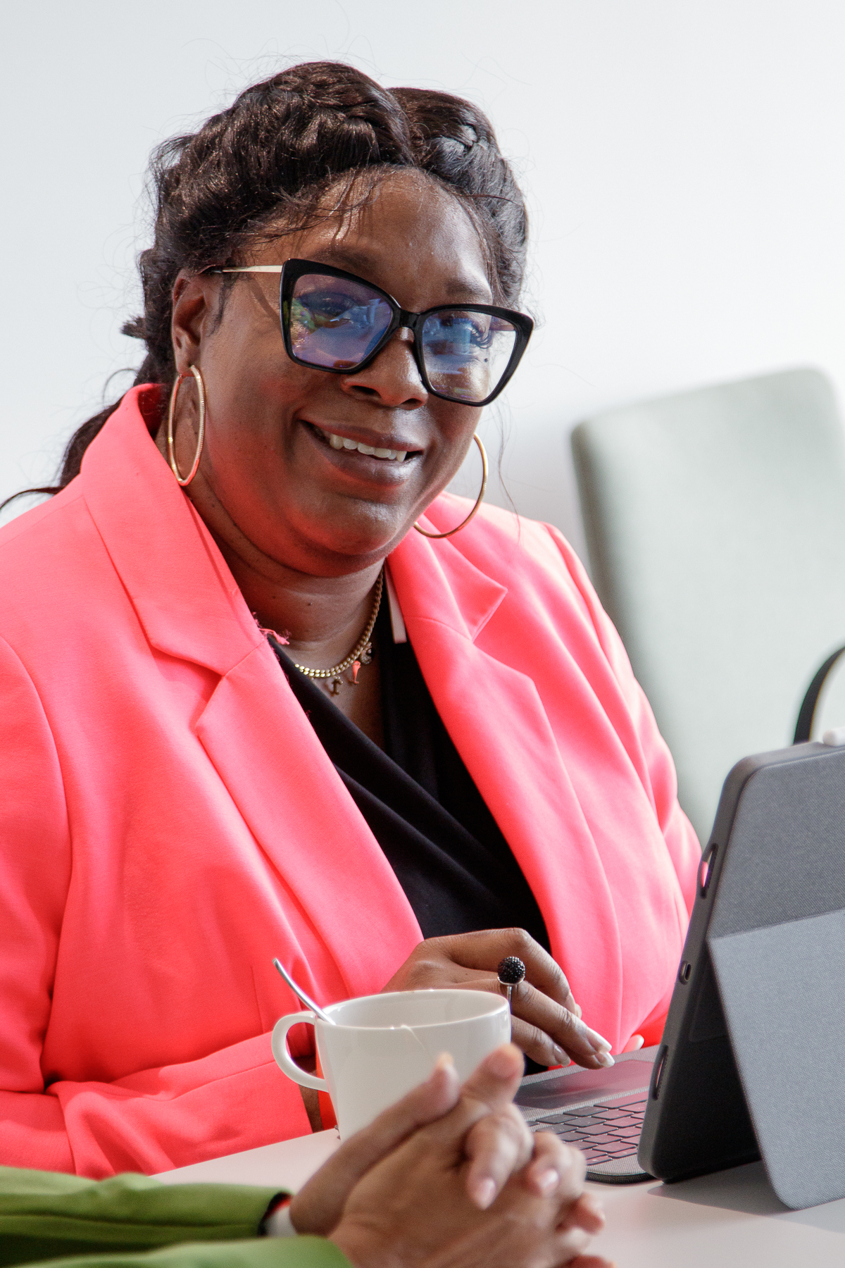 Headshot of Shae, a black woman with glasses. She's sat with her laptop in front of her, wearing a black top and pink blazer.