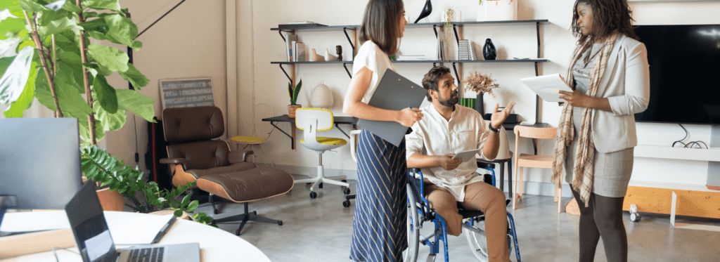 A man with a visible lower-limb difference disability using a manual wheelchair having a workplace meeting with two non-disabled females.