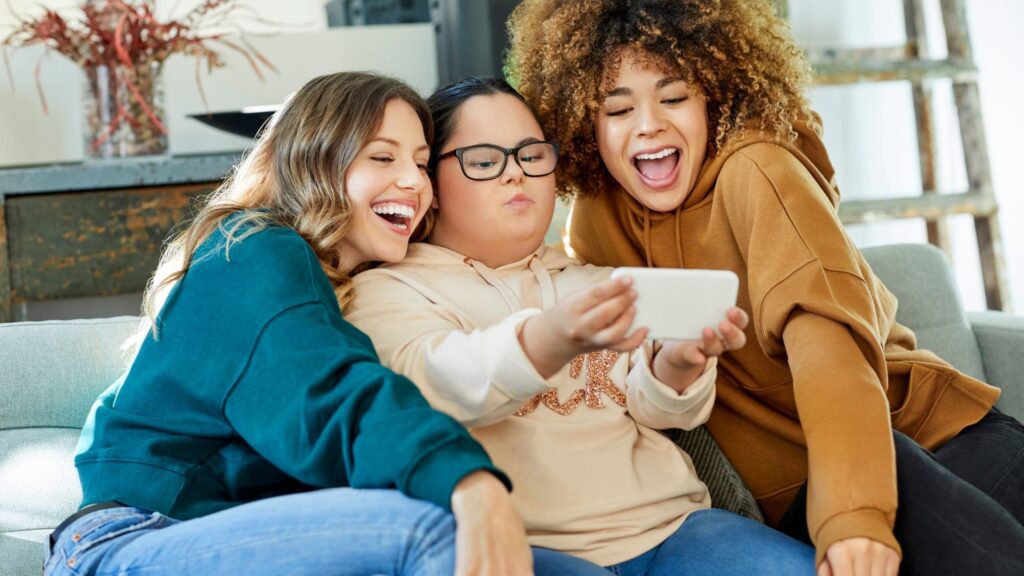 Three women sat on a sofa. The middle woman is holding a phone taking a selfie and the two either side are leaning in, smiling at the camera.