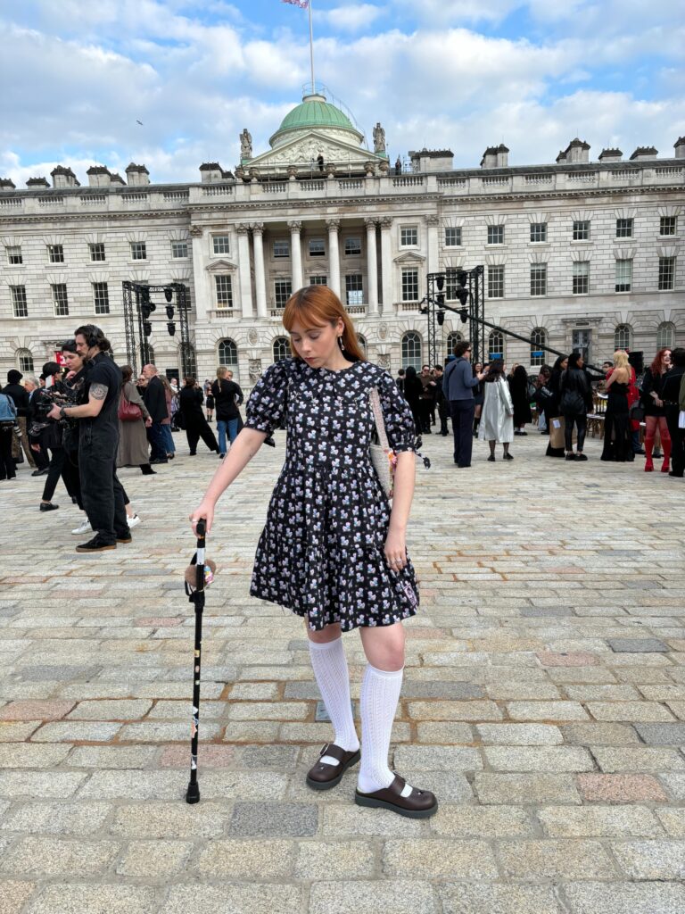 Amy, a white woman with ginger hair, is posing in front of a big building, during a fashion show. She's wearing a printed dress and is holding her walking aid.