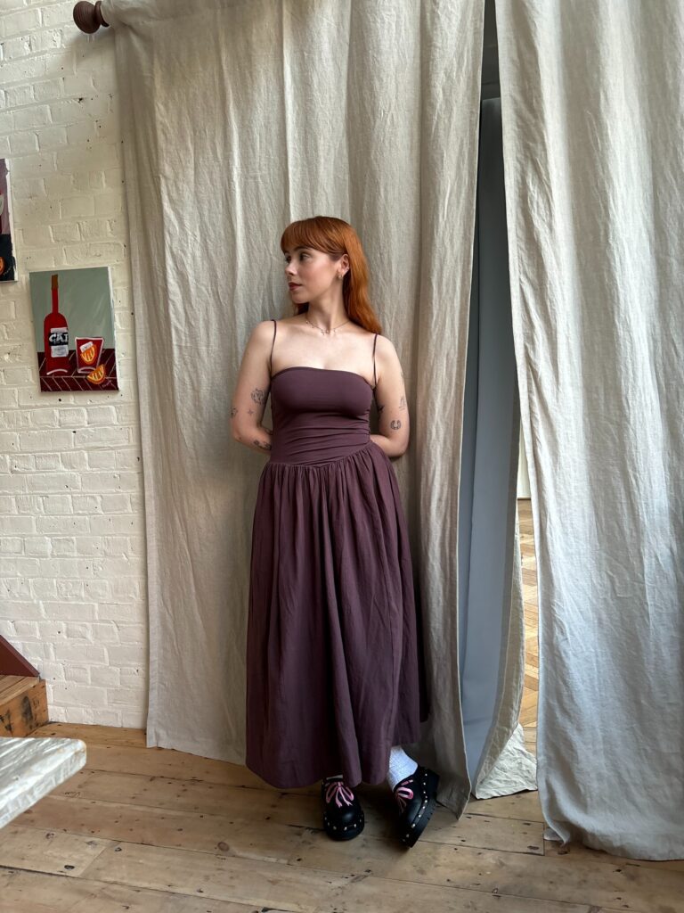 A full body picture of Amy, a white woman with ginger hair, looking to one side, wearing a brown sleeveless dress at an event.