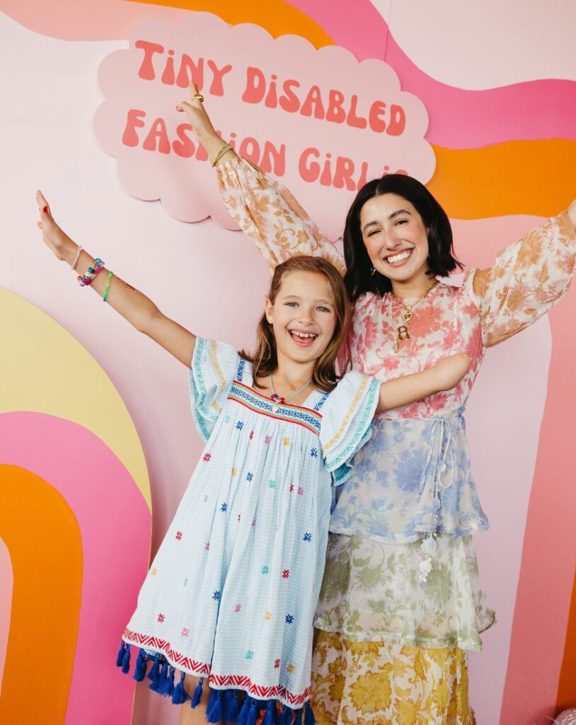 April Lockhart waves with a little girl, both of whom have an upper limb difference. There is a sign behind them that reads: "Tiny Disabled Fashion Girlie."