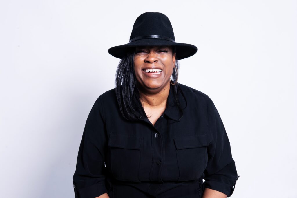 A headshot of Shae, a black woman smiling at the camera, with a black shirt and black hat.