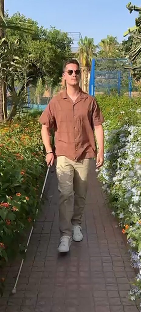 Toby Addison walks through a verdant garden at a TUI resort, using a white cane to guide him.