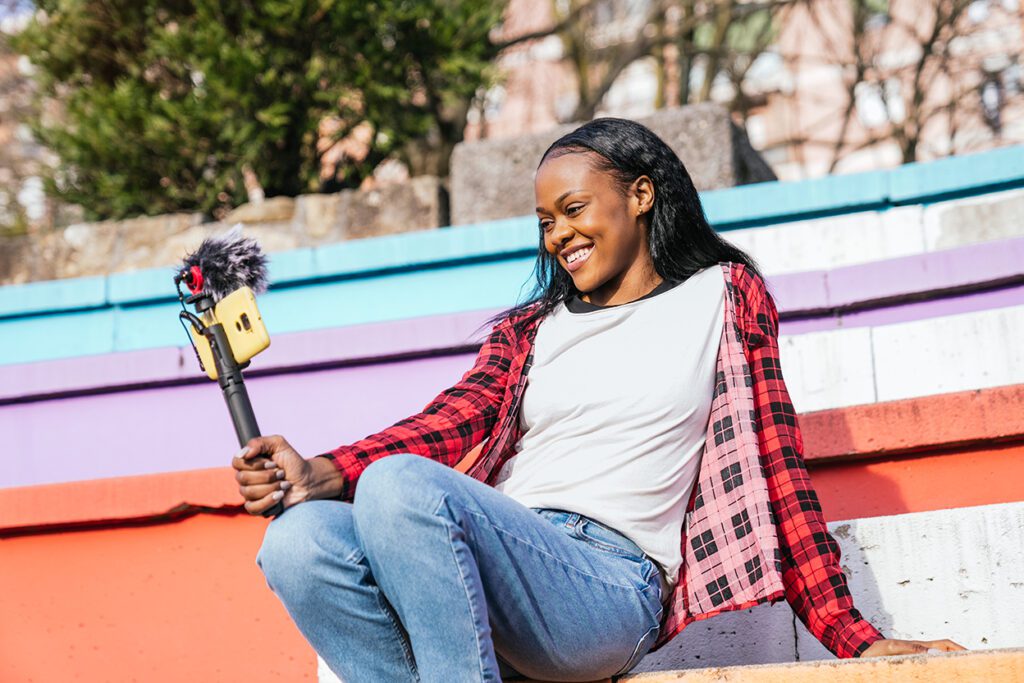 A young woman sits on a set of colourful steps, recording herself on her phone.