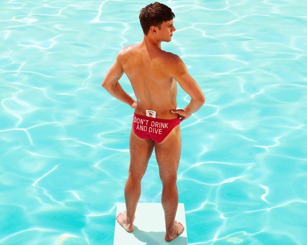 Tom Daley prepares to dive off of a board into a pool. He is wearing knitted swimming trunks with a message on them that reads: "Don't drink and dive."