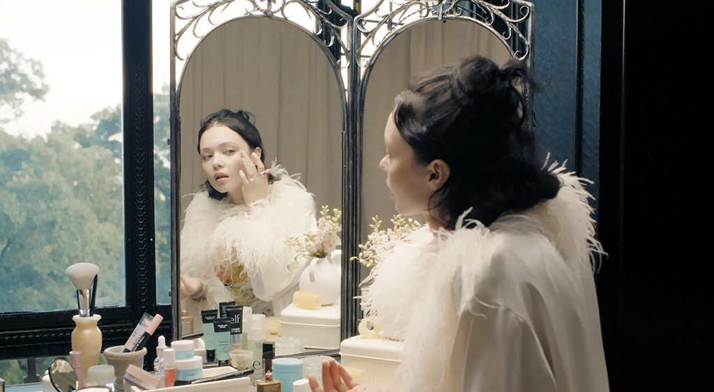 Woman puts on makeup on front of an ornate mirror in a large room as part of an Elf Cosmetics advert.