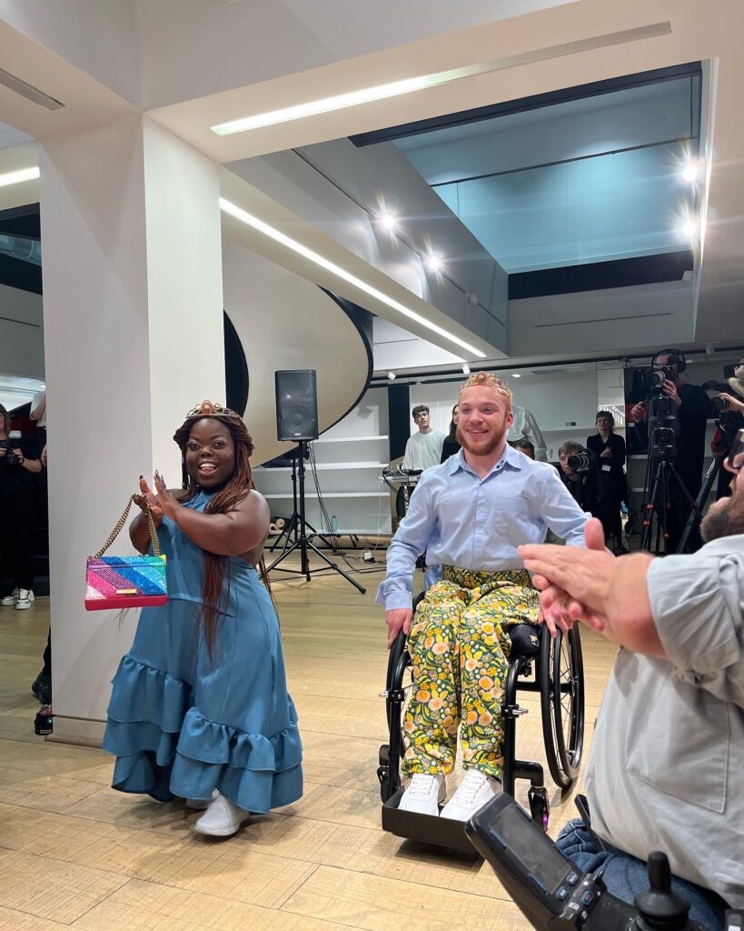 Two models going down the catwalk at an Unhidden fashion show. On the left, a black woman with dwarfism, on the right, a white ginger man on his wheelchair. They're both smiling.