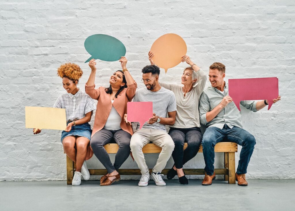 Diverse group of people sitting together, holding illustrative cutout speech bubbles.