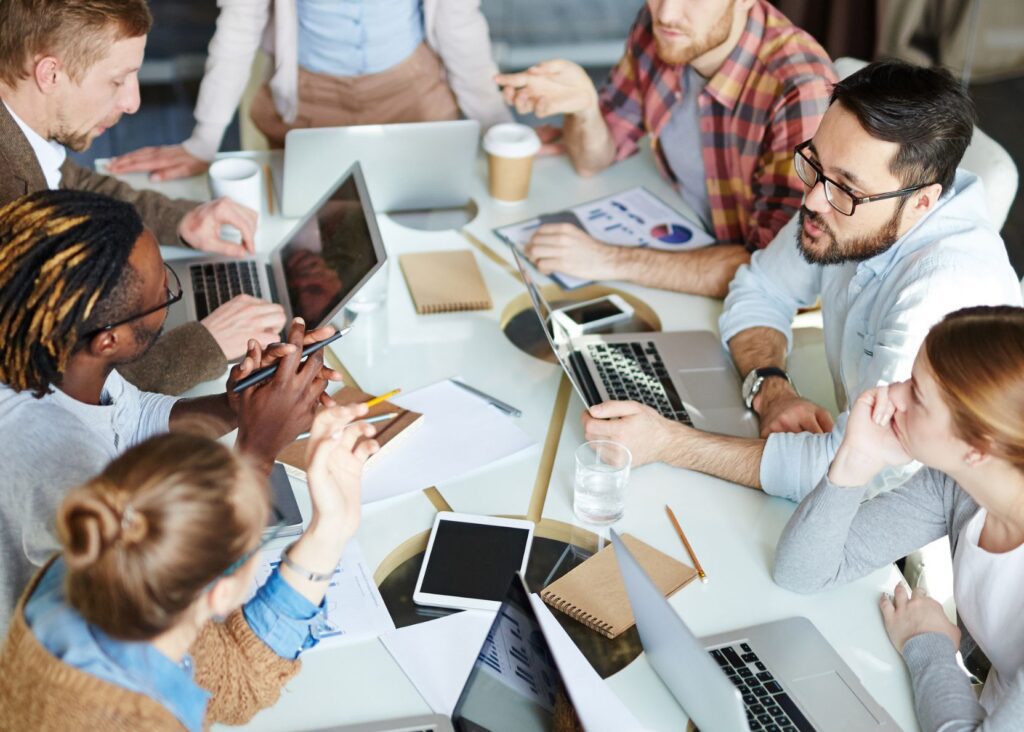 A diverse office team are leaning in for a discussion around a meeting room table.