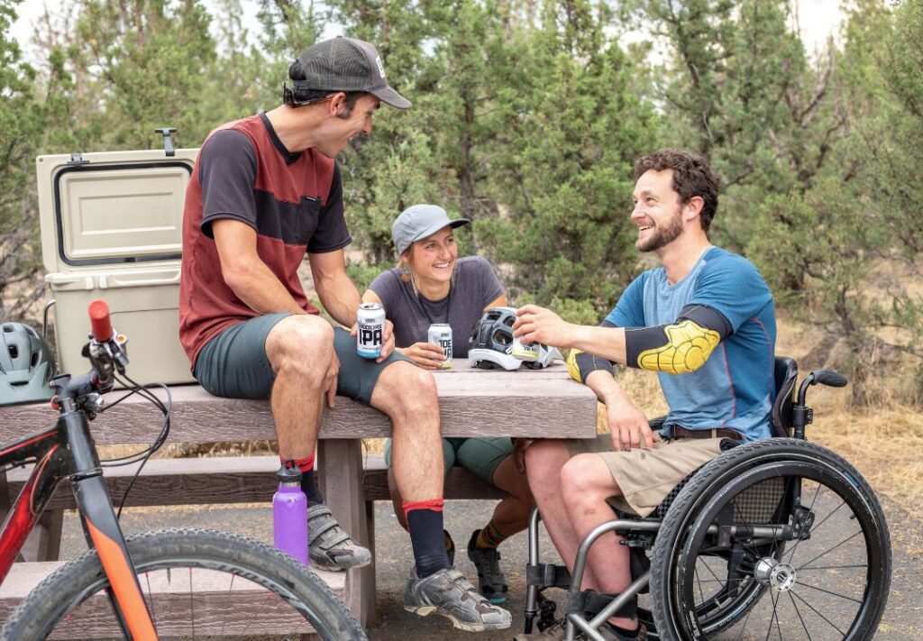 Three people sat together around a park bench. They're smiling at each other. 