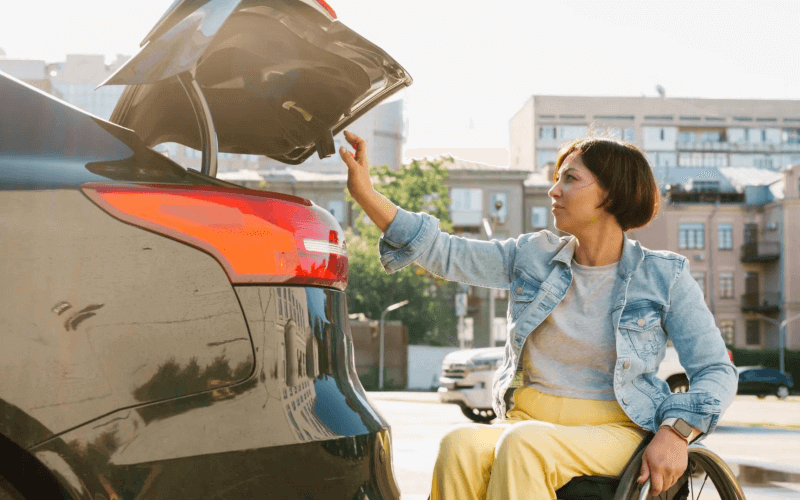 A woman in a wheelchair is opening the boot of their black car. 