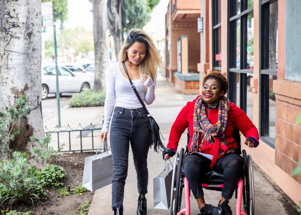 Two friends are walking down a street. One of them is a wheelchair user, and they're both carrying shopping bags.