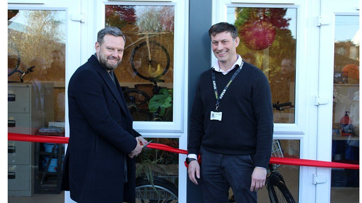 Two white men in front of a bike shop, one of them is cutting a red ribbon in front of it. 