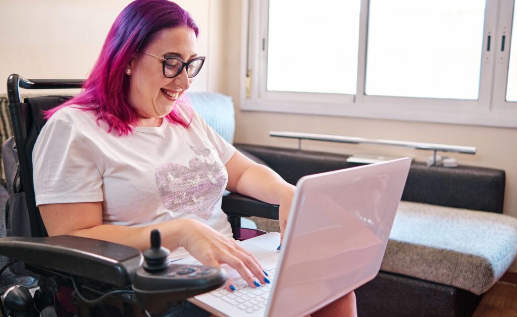 A female powered wheelchair user is smiling as she types on her laptop.